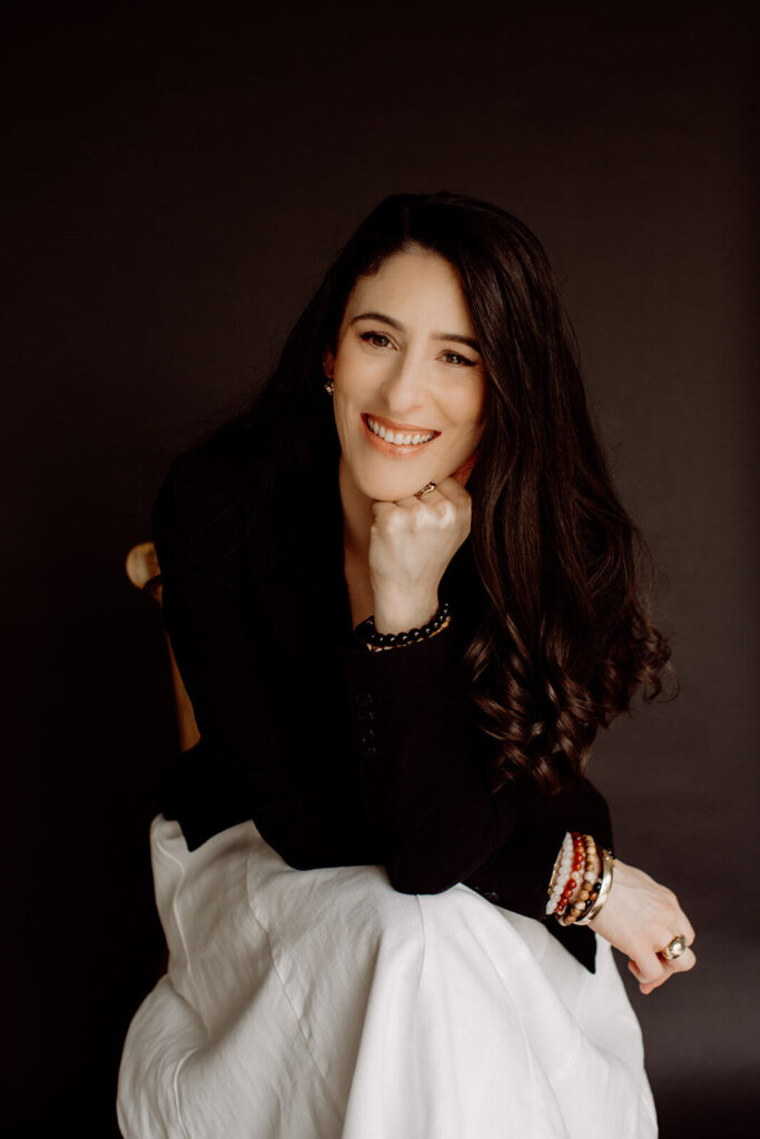 JodiPlumbley_Simmone-11_websize Simmone Helander smiling with her chin resting on her hand, black blazer, dark backdrop