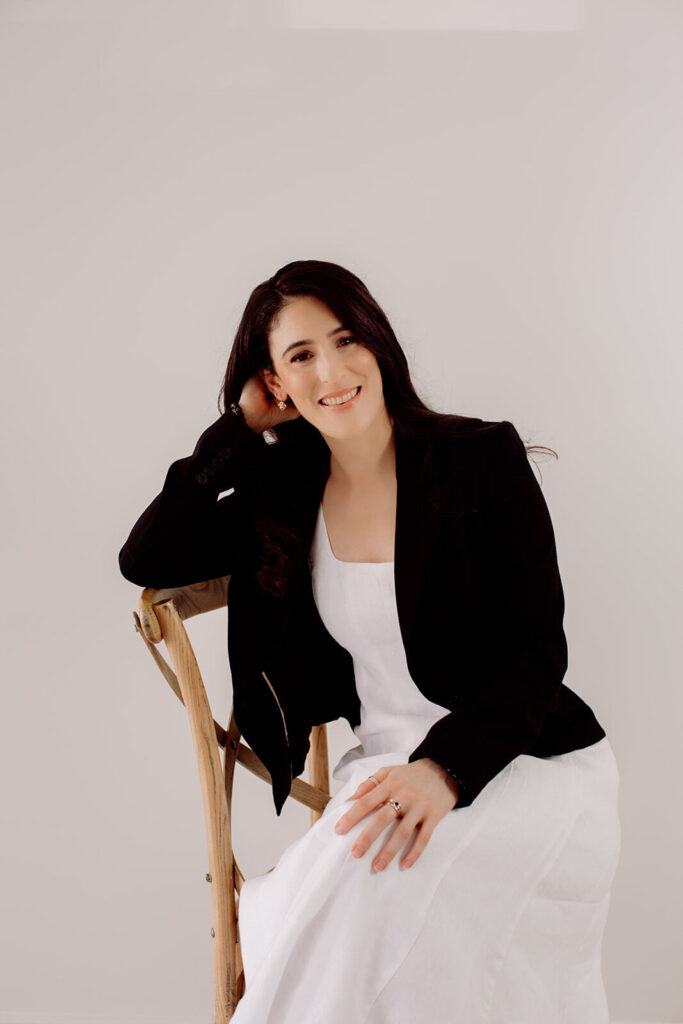 JodiPlumbley_Simmone-15_websize Simmone Helander seated on a timber chair in a black blazer and white dress, smiling