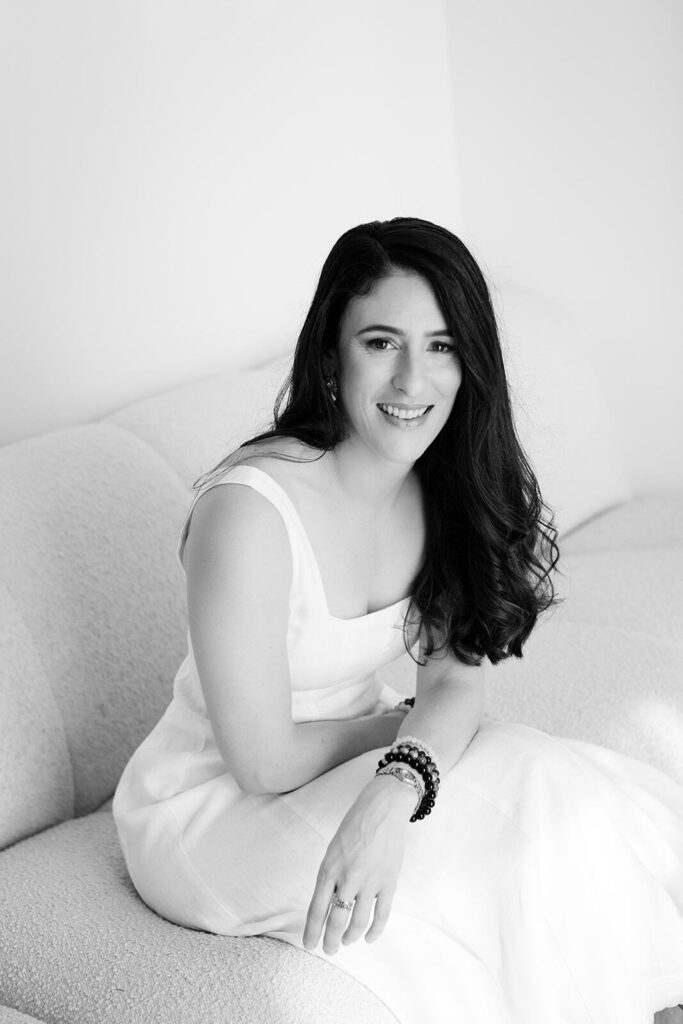JodiPlumbley_Simmone-52_websize Black and white photo of Simmone Helander seated on a couch in a white dress, soft smile