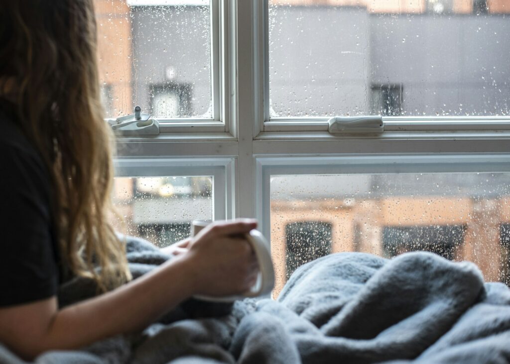 Woman wrapped in a blanket holding a mug by a rainy window