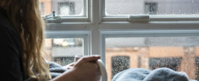 Woman wrapped in a blanket holding a mug by a rainy window